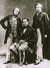  Three middle-aged men, with the one in the middle sitting down. All wear long jackets, and the shorter man on the left has a beard.