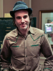 A smiling man wearing a gray hat with piping above the band, and a tan Western style shirt, stands in an office, posing for the camera.