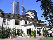 Un edificio neoclásico de dos pisos que muestra influencias de la arquitectura japonesa, con una torre central de dos plantas. En el primer plano es un jardín y una pista de tenis y en el fondo son los rascacielos.