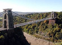 Ponte de suspensão entre duas torres de tijolos construído, ao longo de um desfiladeiro arborizado, mostrando lama e água na parte inferior. Na distância são colinas.