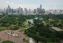 Um parque com muitas árvores e um lago; um bronze estátua estar em frente ao parque; muitos edifícios no fundo