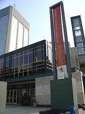 O Francis Winspear Centre for musical com uma bandeira na frente para a torre Edmonton Symphony Orchestra e Century Place por trás dele.